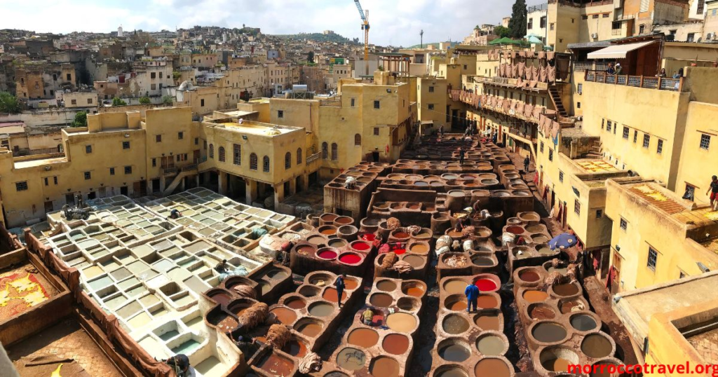 A vibrant scene of Casablanca, capturing its essence as a captivating travel destination. In the foreground, a bustling local market with colorful stalls offering spices, textiles, and artisanal crafts, surrounded by travelers in modest casual clothing. The middle ground features the iconic Hassan II Mosque, showcasing its stunning architecture with intricate tile work, set against a bright blue sky. In the background, the ocean waves gently lap at the shore, reflecting the sunlight. The lighting is warm and inviting, suggesting an early morning or late afternoon. The mood is lively and welcoming, symbolizing the charm of budget-friendly exploration in Morocco. The viewpoint should be slightly elevated, offering a panoramic perspective of this captivating locale.